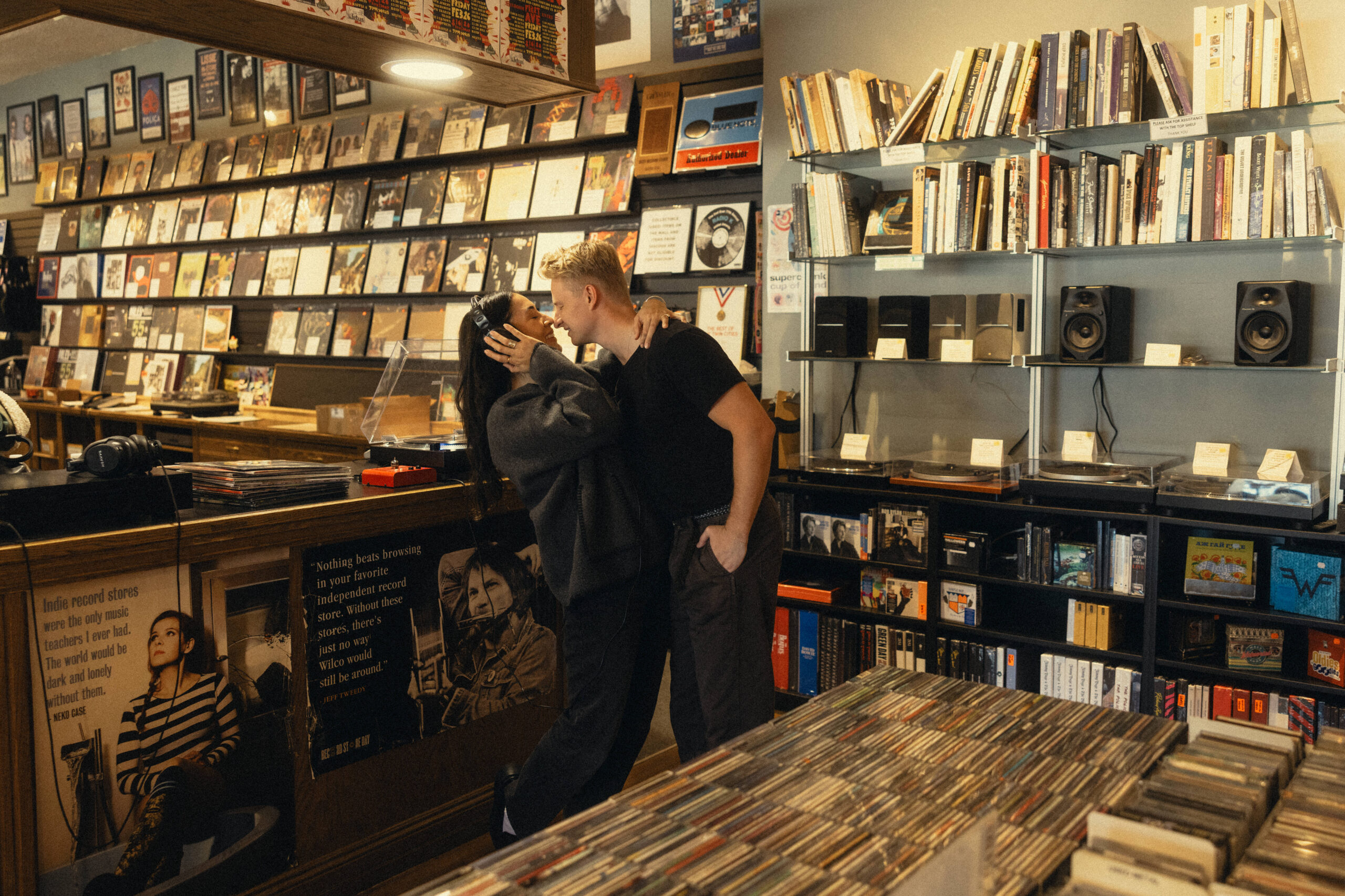 a young couple in a record store. the girl is wearing head phones listening to a record with her arm around her fiance